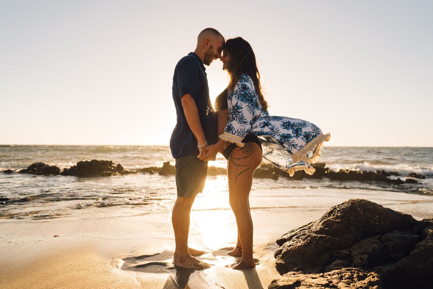 A silhouetted couple shares an intimate moment on the beach at sunset with waves in background.