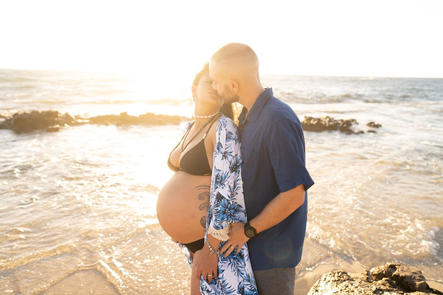 A maternity photoshoot at sunset features a couple embracing on a rocky beach with golden light.