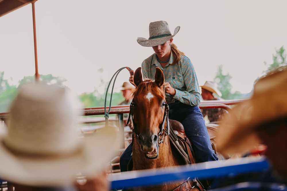 A cowgirl in a hat rides a chestnut horse at a rodeo arena, holding reins with spectators in cowboy hats nearby.