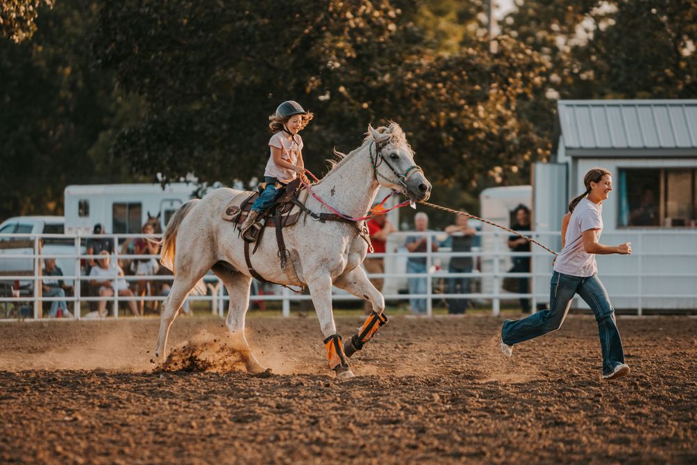 Rider on a white horse in an arena while another person runs alongside.