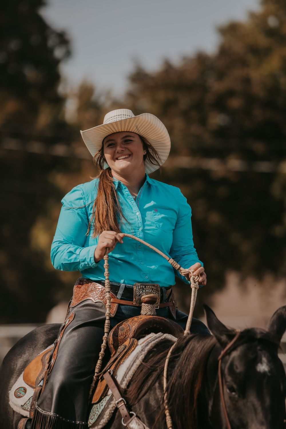 A rider in a bright turquoise shirt and cowboy hat guides a horse during an equestrian event.