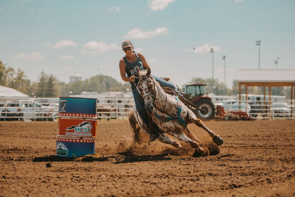 A barrel racer leans into a sharp turn around a blue barrel in a dusty arena.