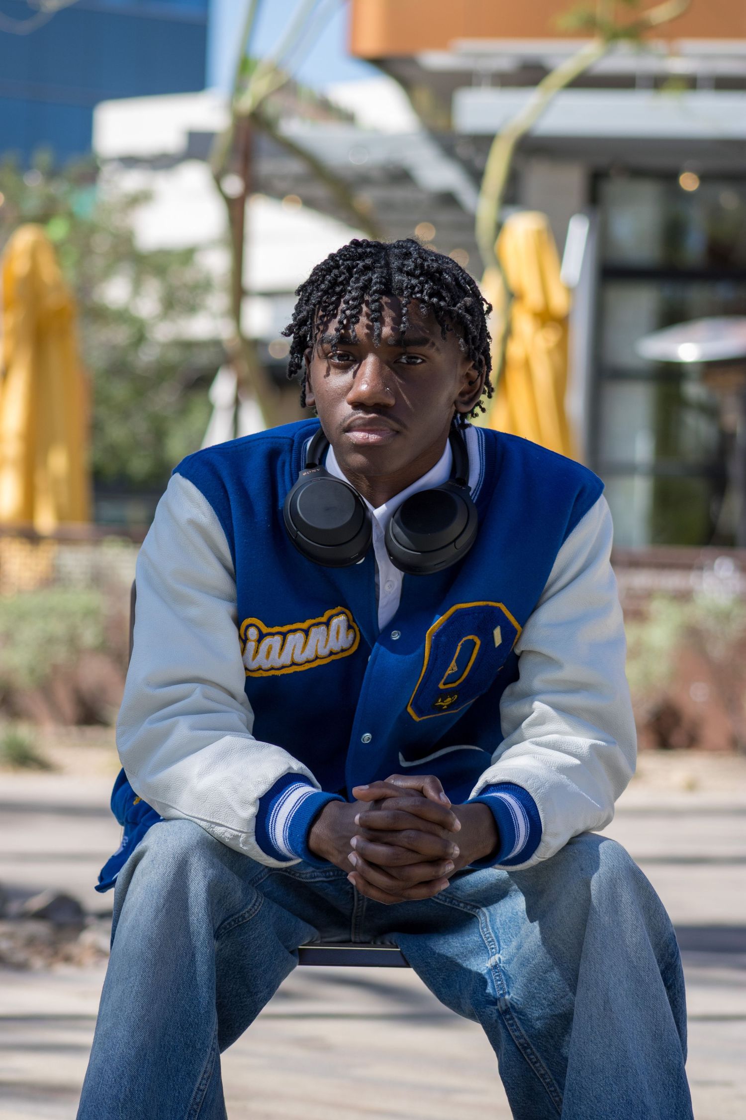 Young man in a blue varsity jacket with headphones sitting outdoors looking serious.