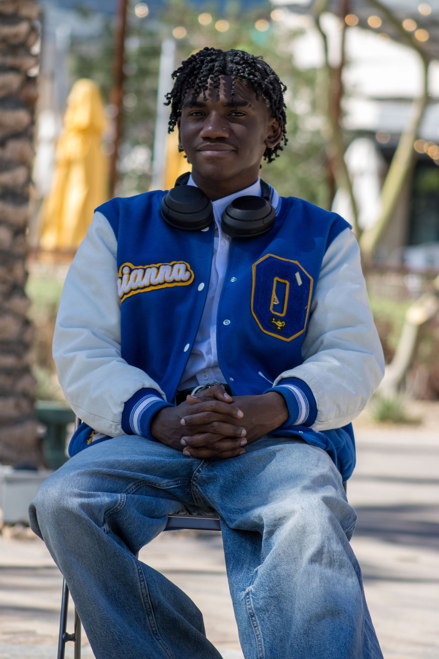 Young man in blue Olathe Pirates varsity jacket seated outdoors with headphones around neck.