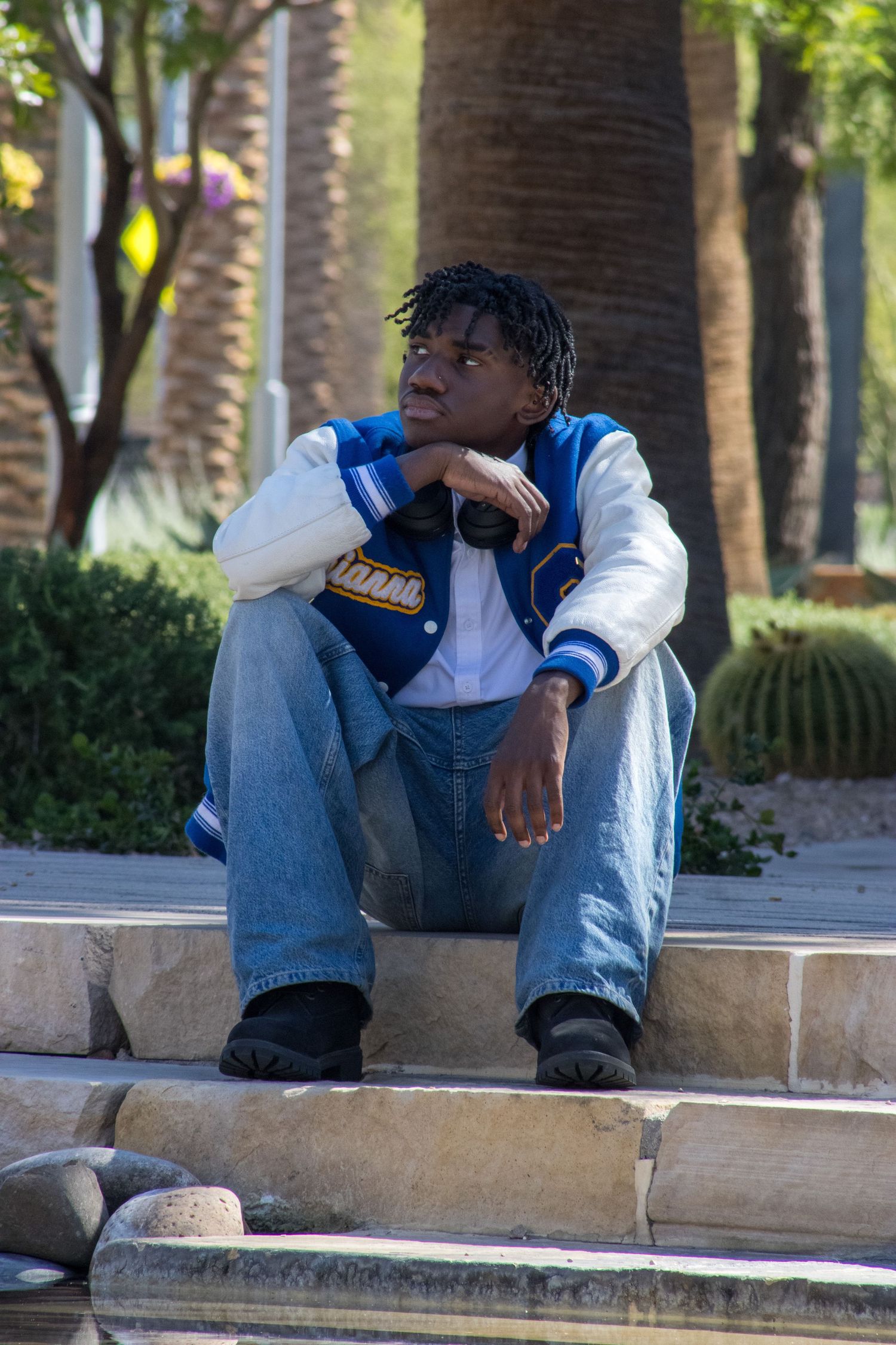 Teen in blue varsity jacket sitting on stone steps outdoors looking thoughtfully to the side.