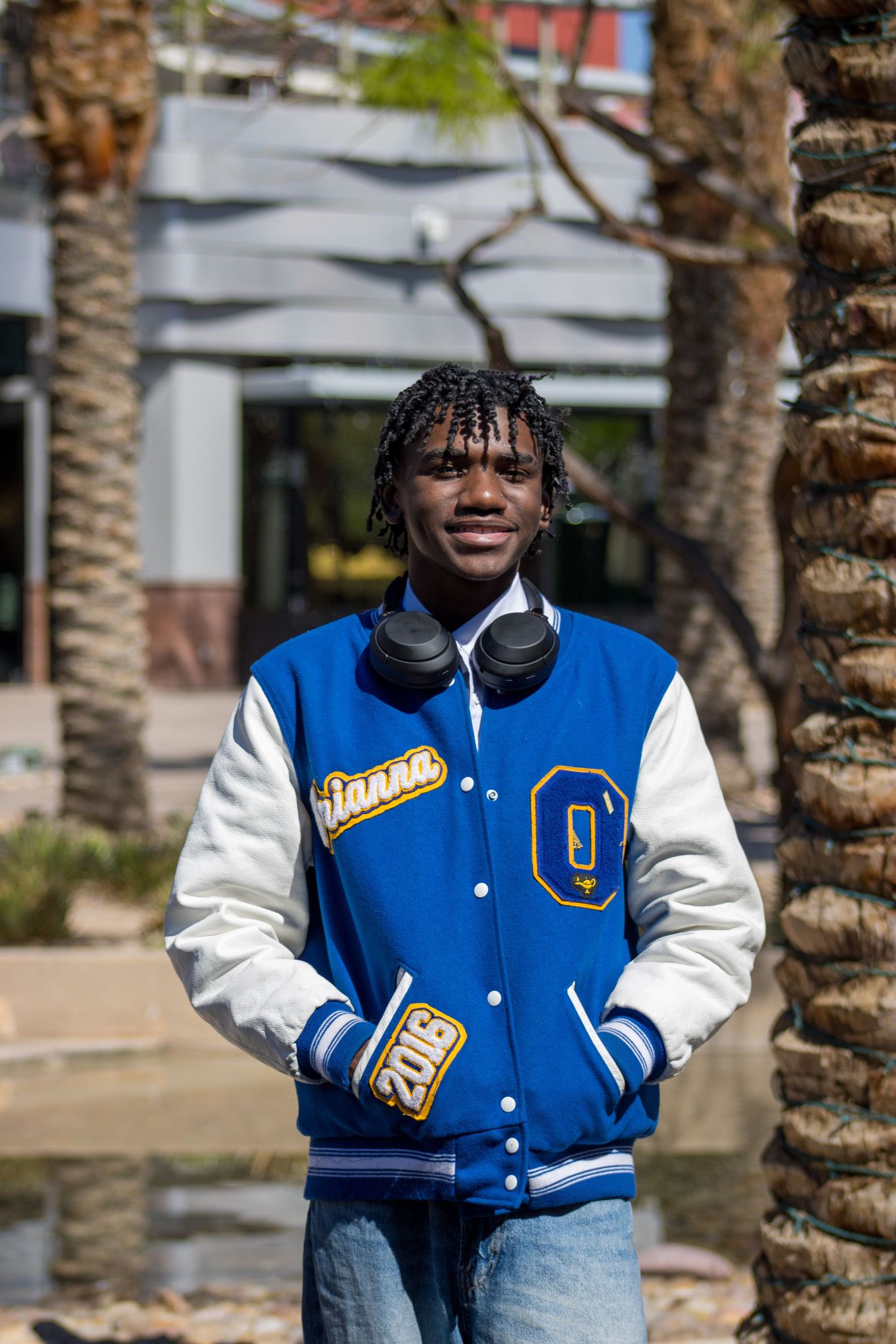 Young man proudly wearing blue Olathe Pirates letterman jacket standing near palm trees outdoors.