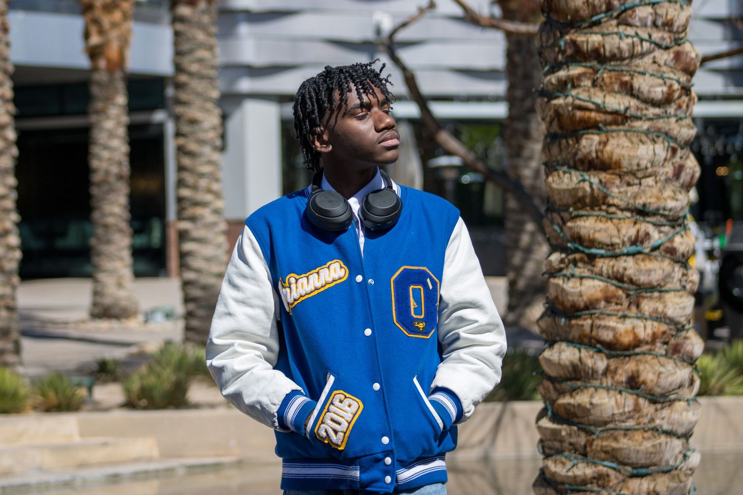 Teen in blue and white varsity jacket standing by palm tree in sunny outdoor setting.