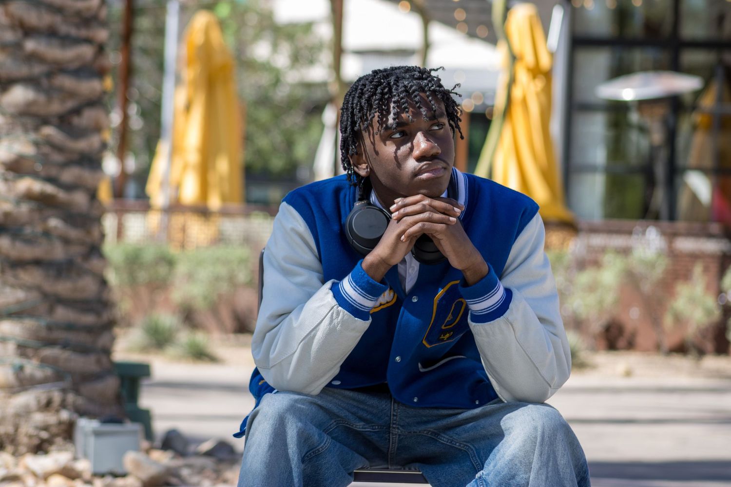 Young man in blue varsity jacket sitting outdoors with hands clasped, looking downward pensively.