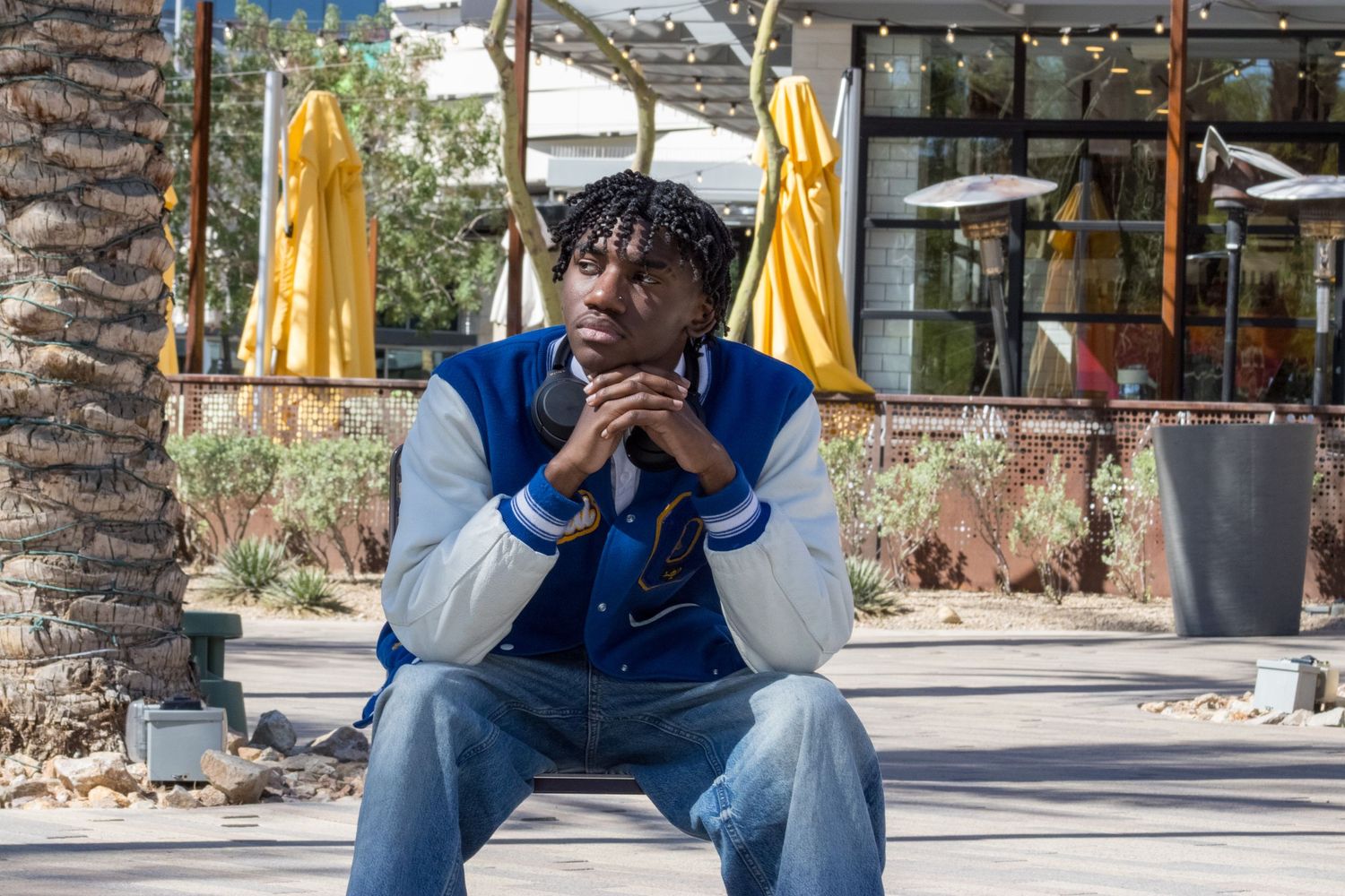 Teen in blue letterman jacket seated outdoors with hands clasped near yellow umbrellas background.