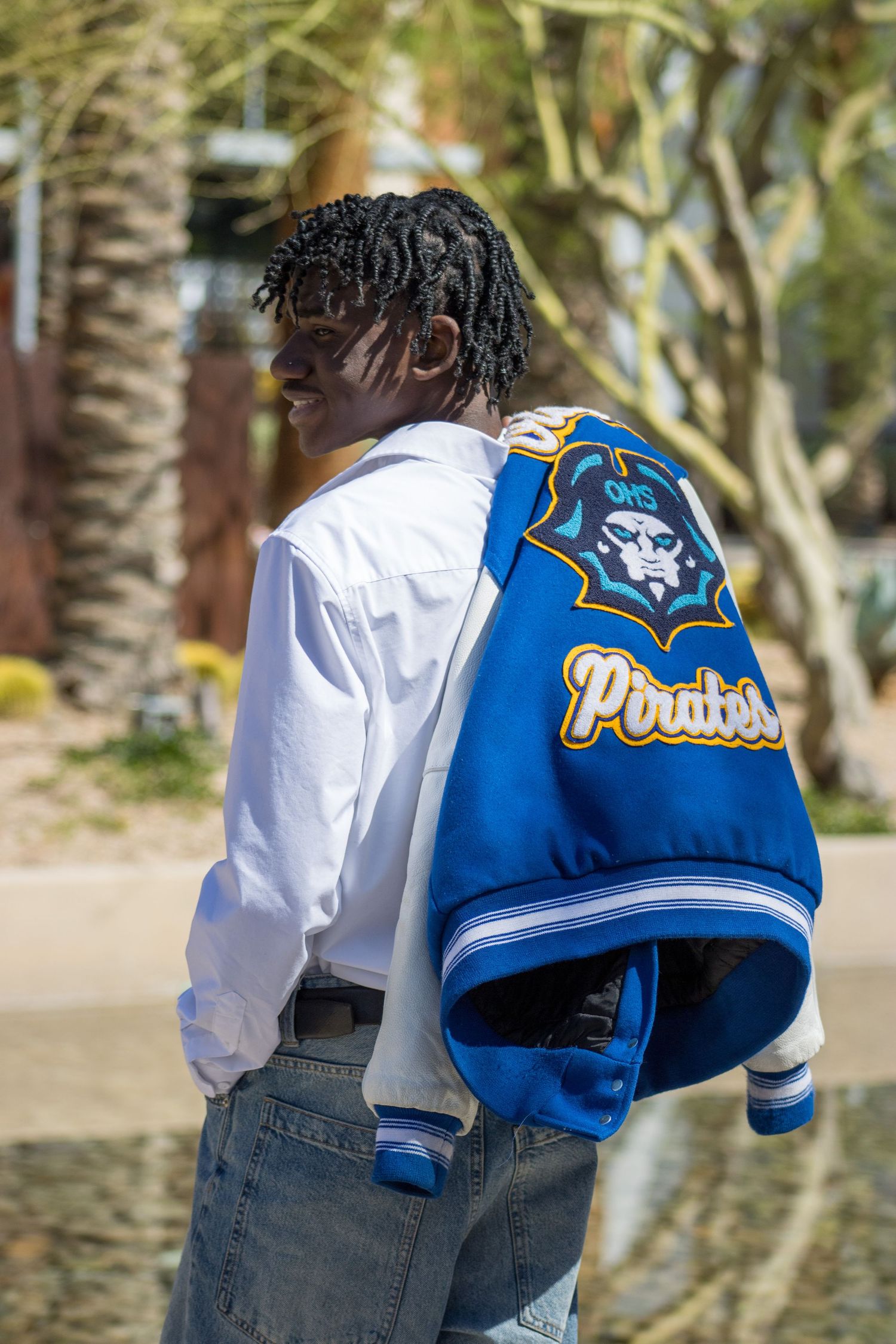 Teen smiling while draping blue Olathe Pirates varsity jacket over shoulder outdoors.