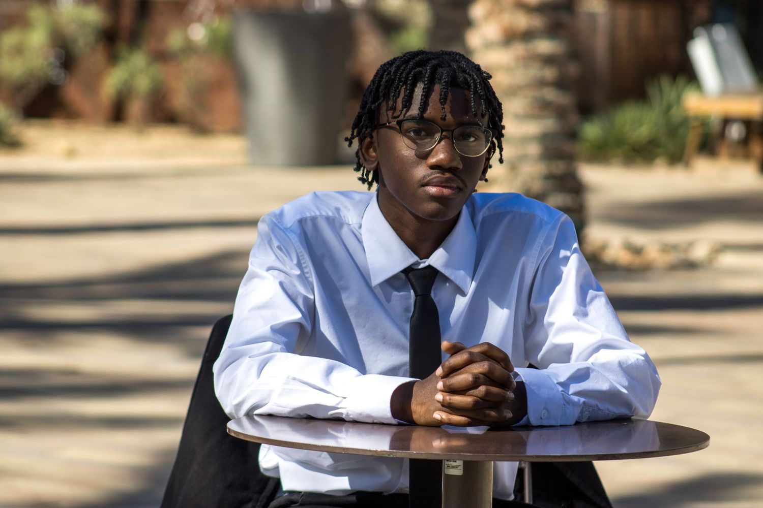 Young man in dress shirt and tie seated at outdoor table with hands clasped forward.