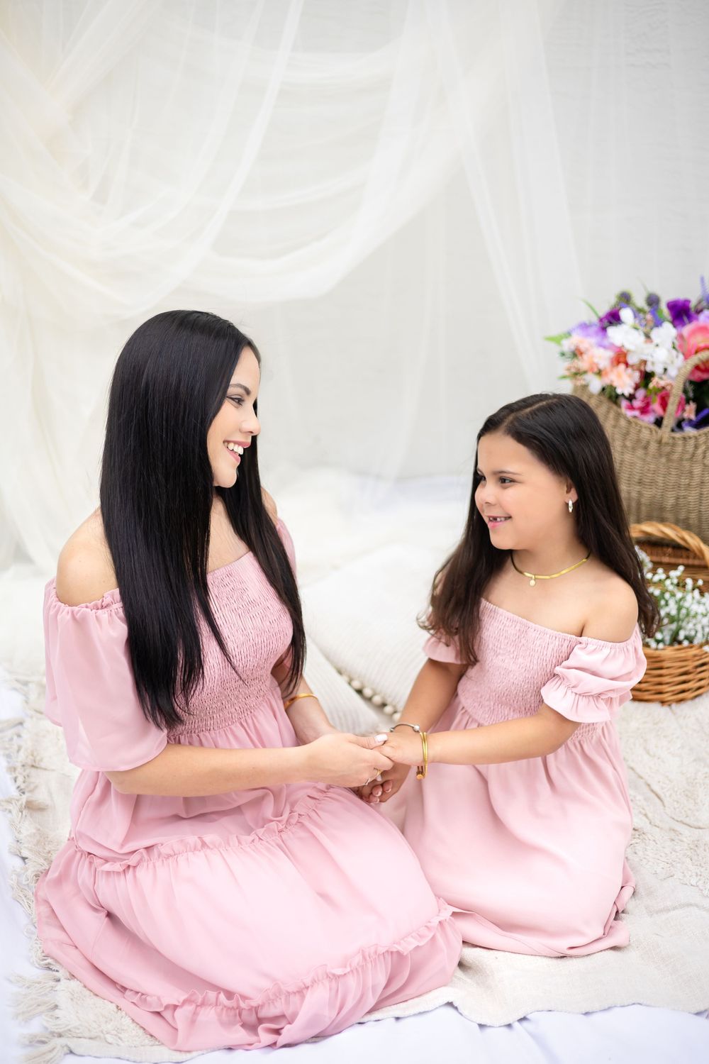Two people in matching pink dresses share an intimate moment on white bedding with floral decorations.