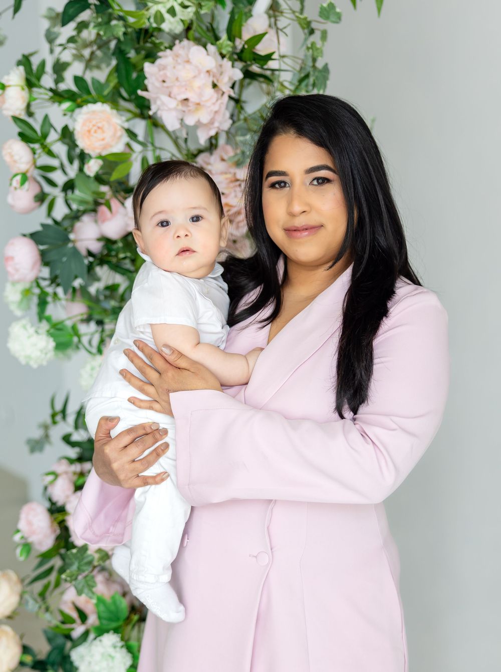 Floral backdrop frames mother and baby wearing matching pink and white outfits for a spring portrait session.