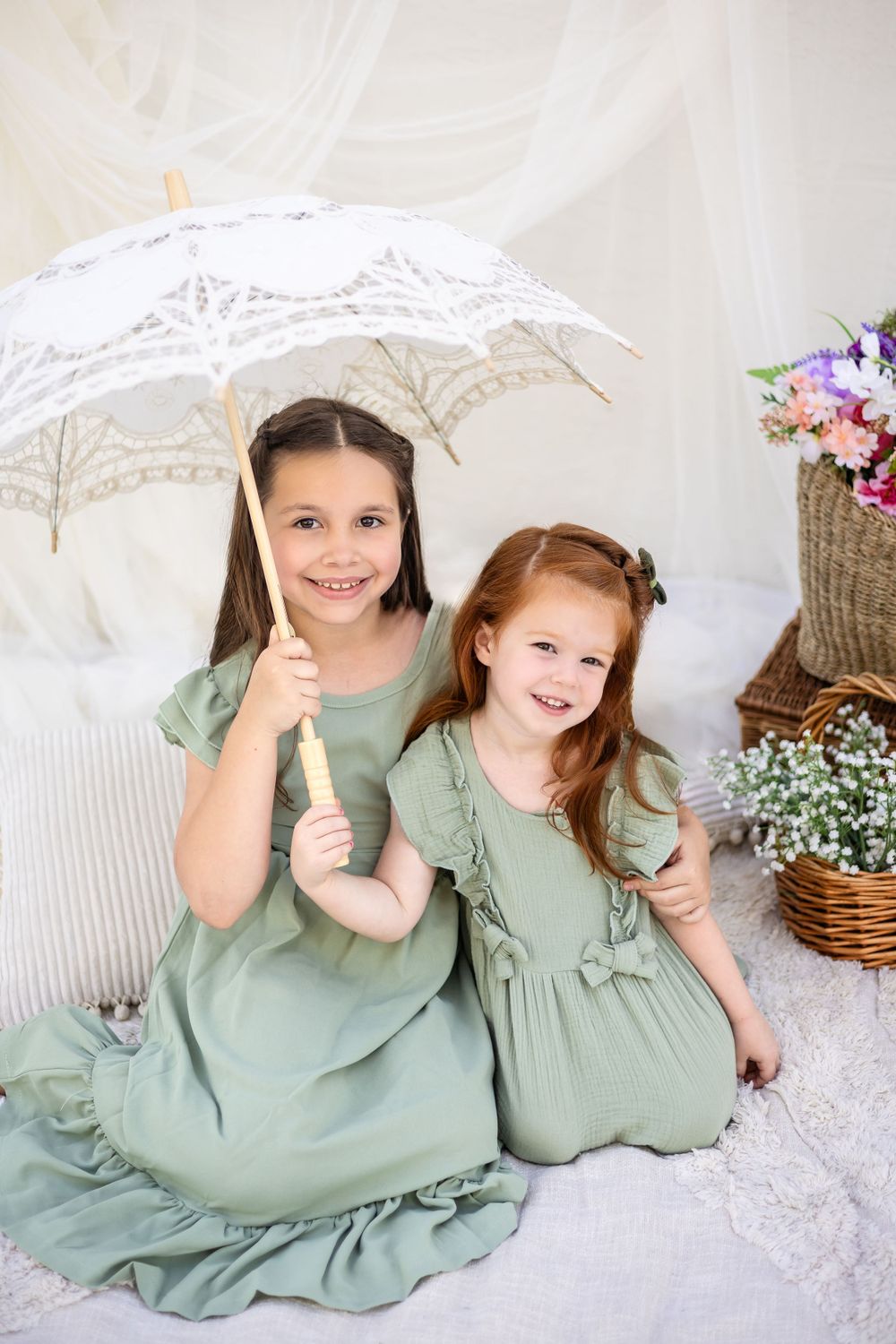 Two children in matching sage green dresses pose with lace umbrella against white backdrop.