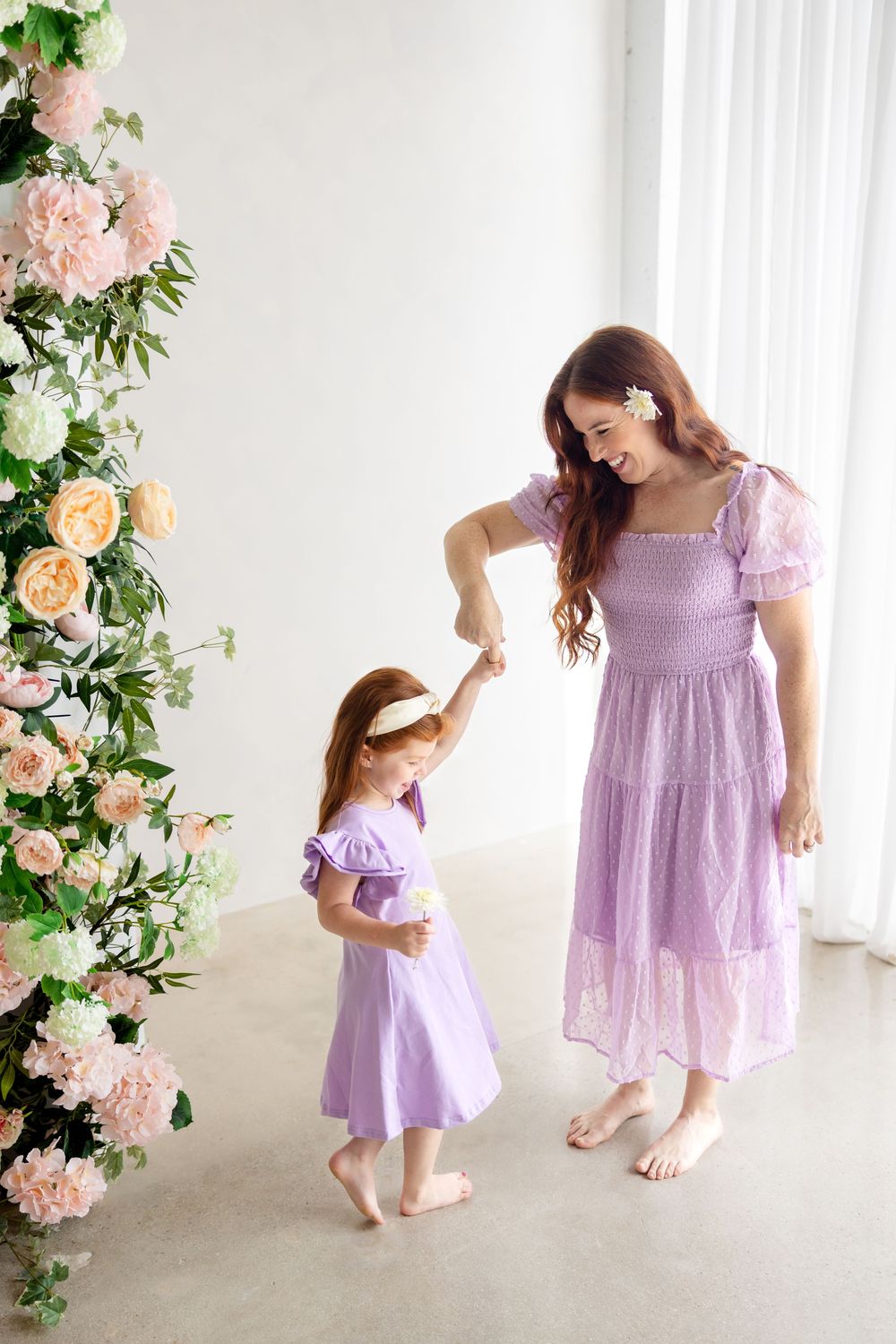 Mother and daughter wearing matching lilac dresses dance together beside a floral arch with pink and peach roses.