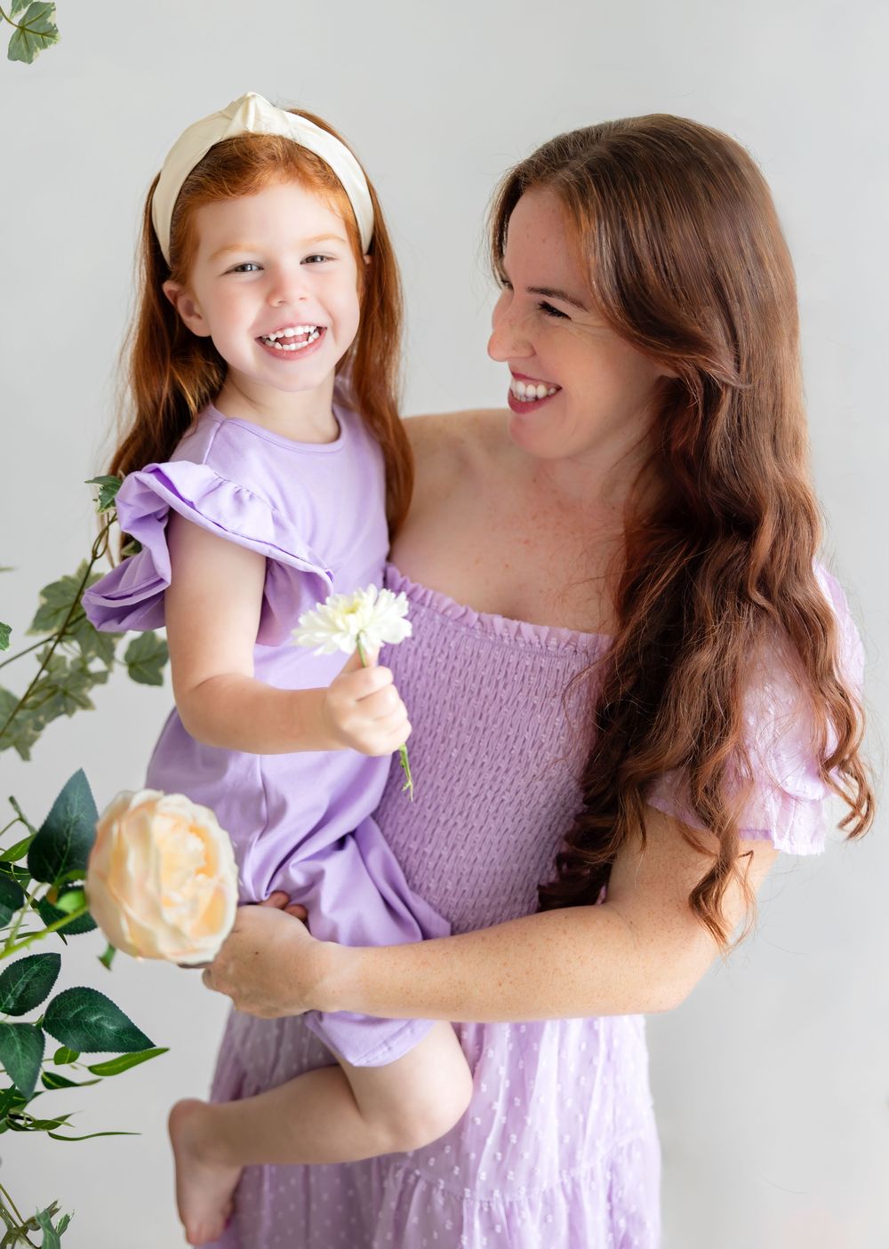 Two people in lilac dresses sharing a joyful moment with flowers against a white backdrop.