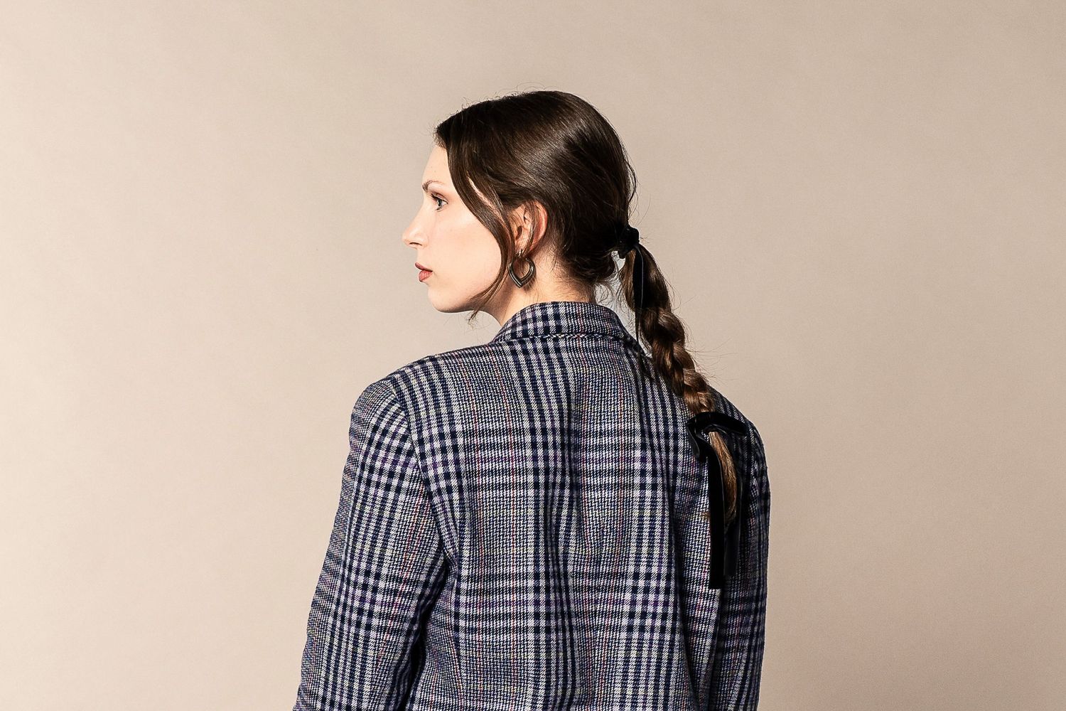 Back view of person wearing plaid blazer with long braided dark hair against beige background.