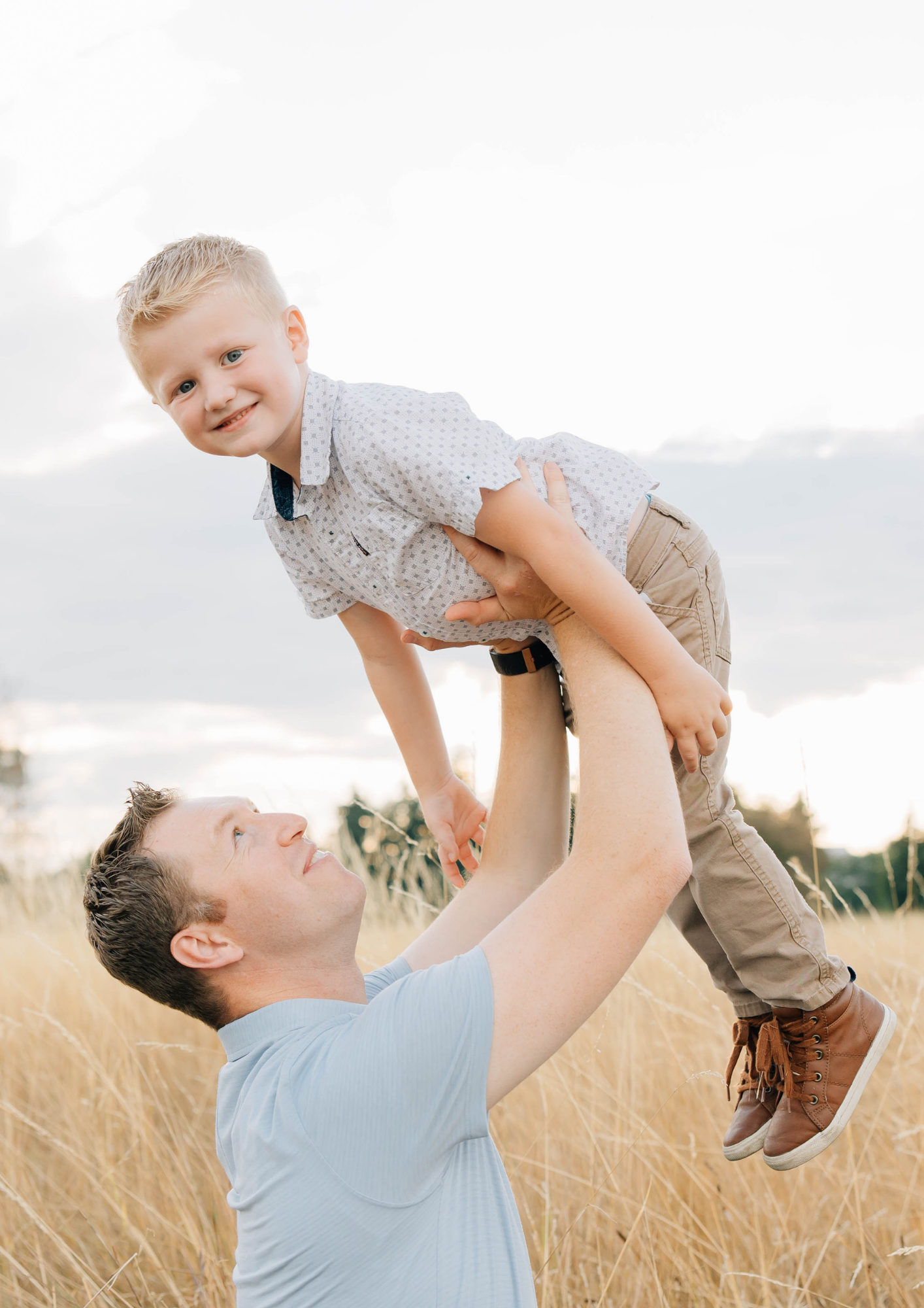 Father lifting smiling young boy overhead in golden grass field outdoors.