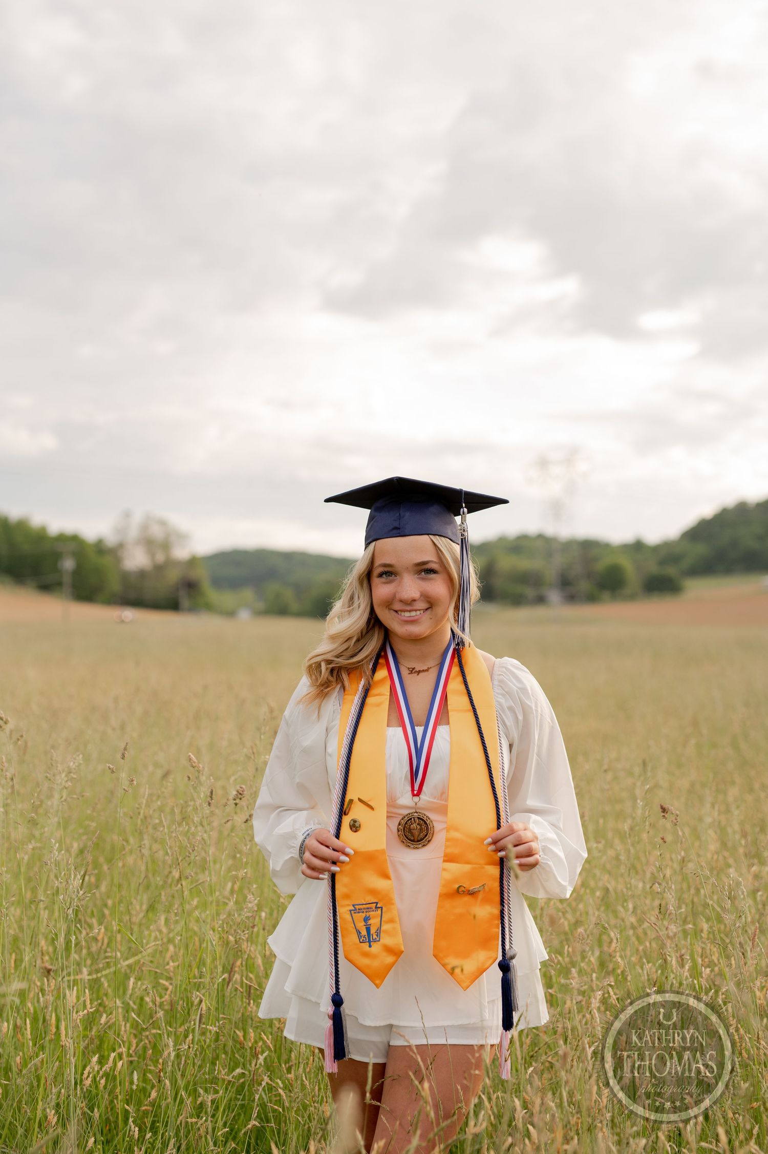 Graduate in academic regalia and gold honors stole stands in field of tall grass.