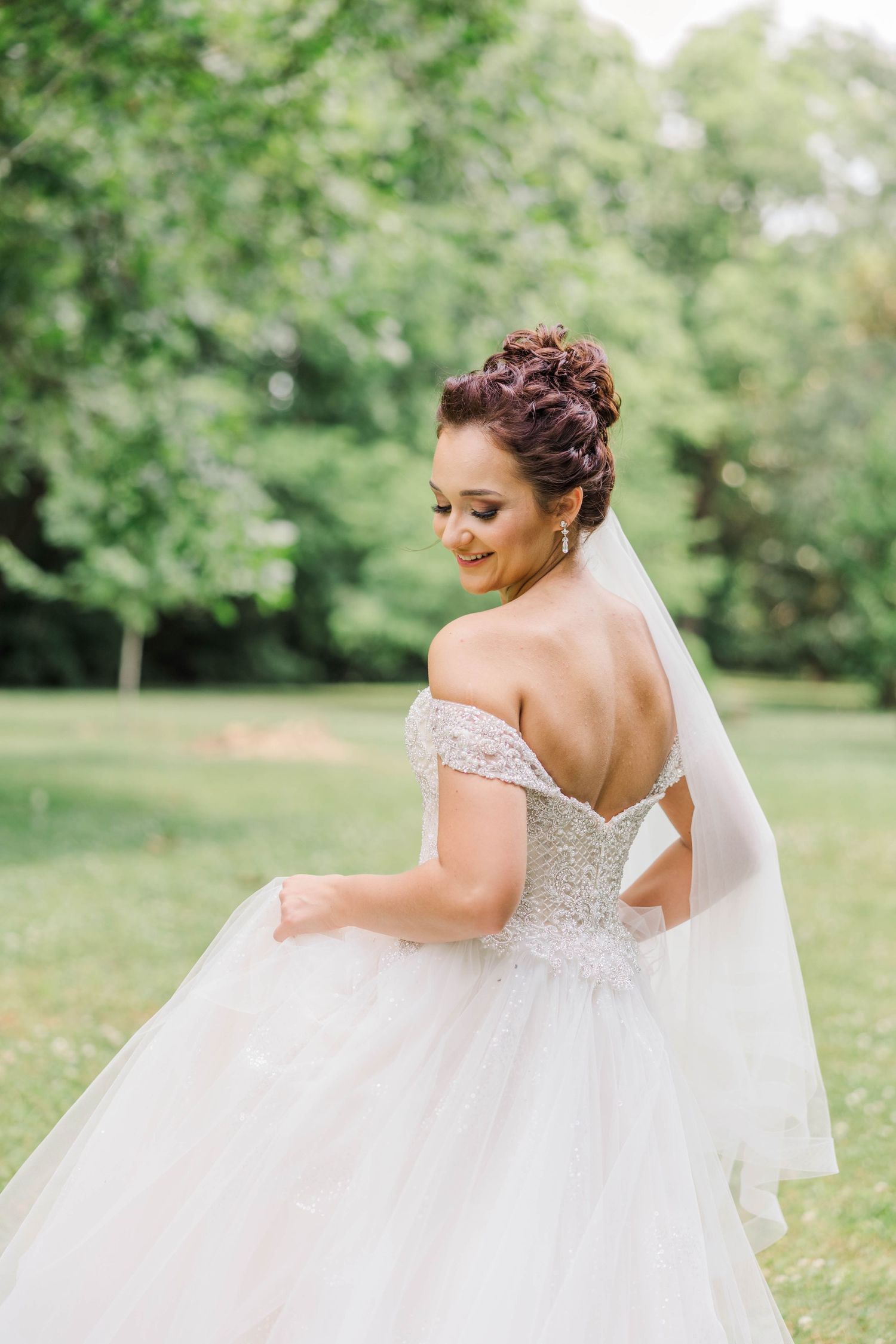 Romantic back view of bride in off-shoulder ballgown with veil in garden setting.