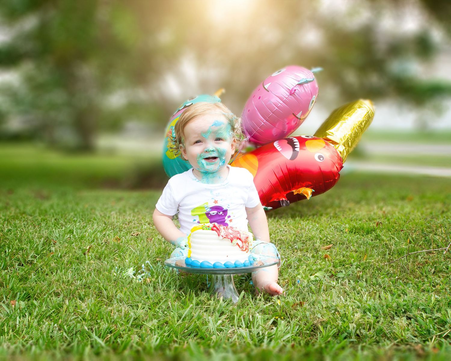 A toddler sits in green grass with colorful heart-shaped balloons during a sunny outdoor photoshoot.
