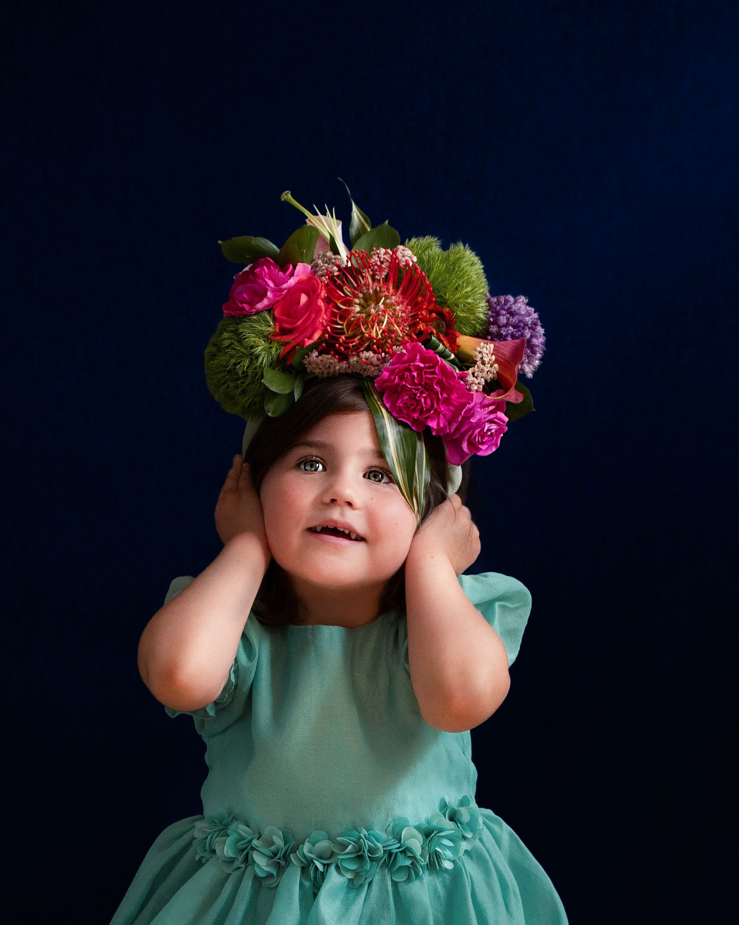 Small child in turquoise dress with vibrant floral crown poses against dark background. Frida Kahlo style