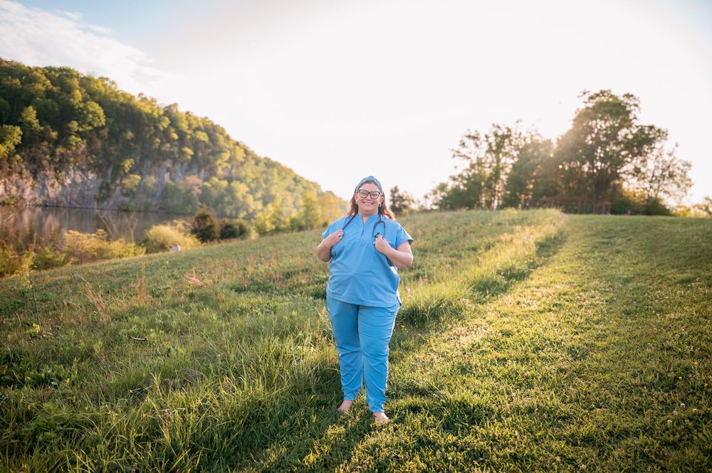 Healthcare worker in blue scrubs walking through a sunlit green meadow with rolling hills in the background.