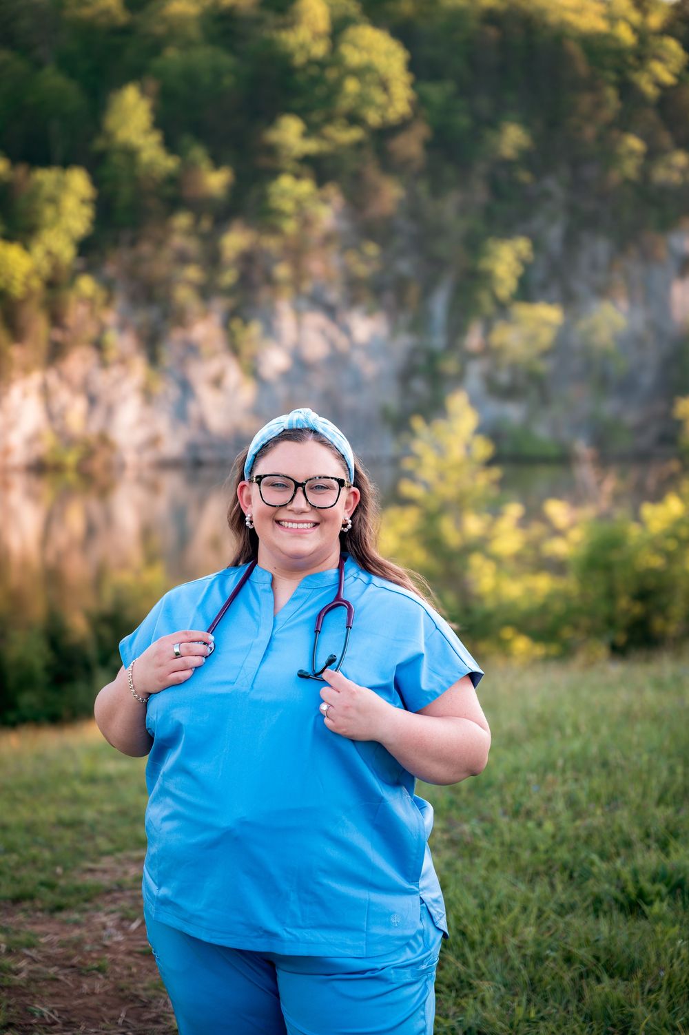 Smiling female nurse in blue scrubs with stethoscope poses outdoors near a scenic lake and rock cliff.