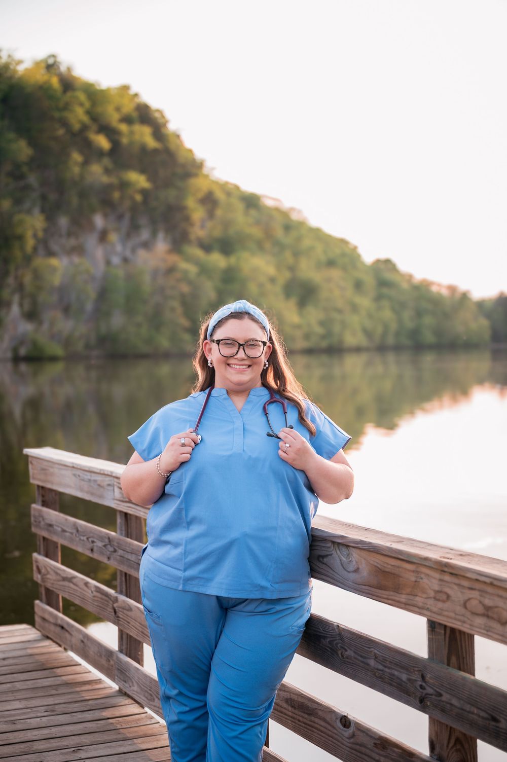 Smiling nurse in blue scrubs with stethoscope poses on wooden dock by calm lake surrounded by trees.