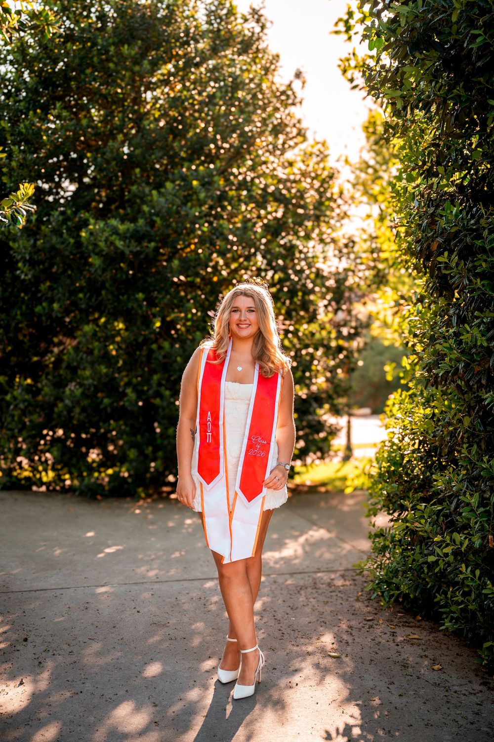 Young woman in white dress and red graduation stole smiles on a tree-lined path during golden hour.