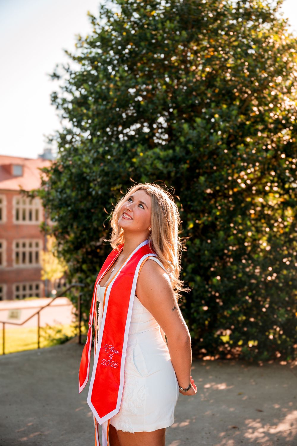 Smiling blonde woman in white dress and red graduation stole posing outdoors near green trees.