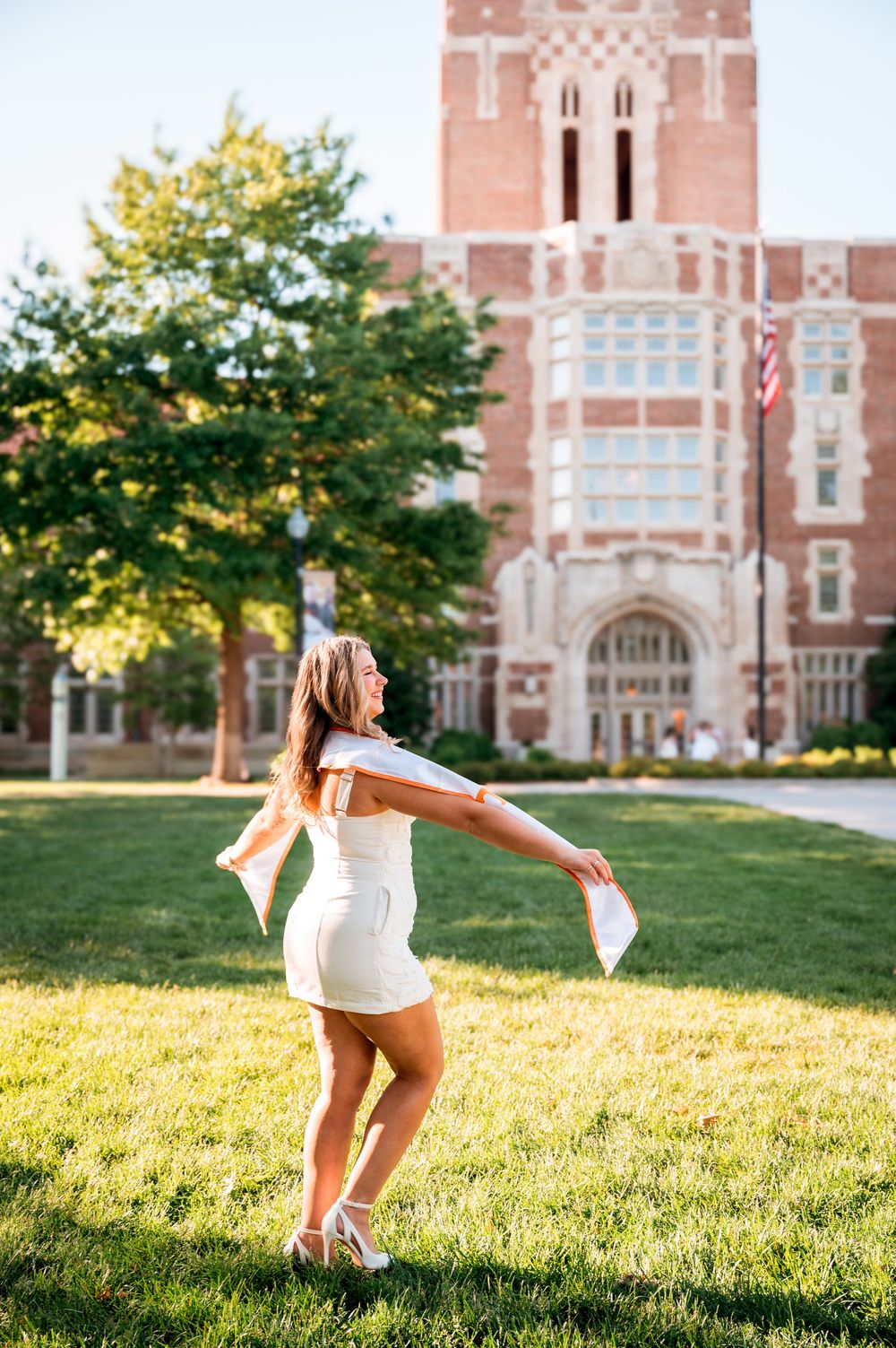 A young woman in a white dress twirls joyfully on a green lawn in front of a brick university building.