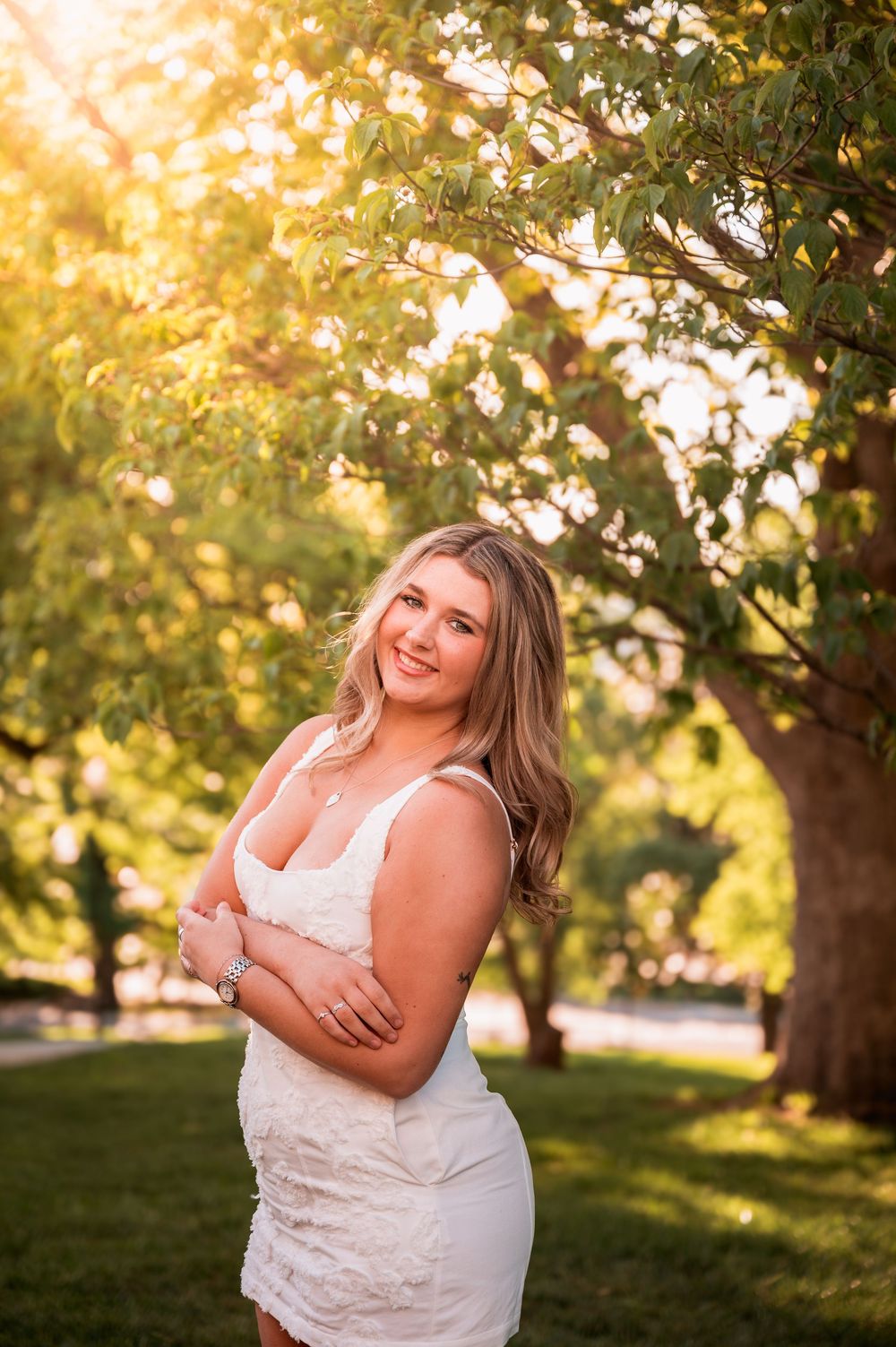 Smiling young woman in white dress poses outdoors under golden sunlight with lush green trees in background.