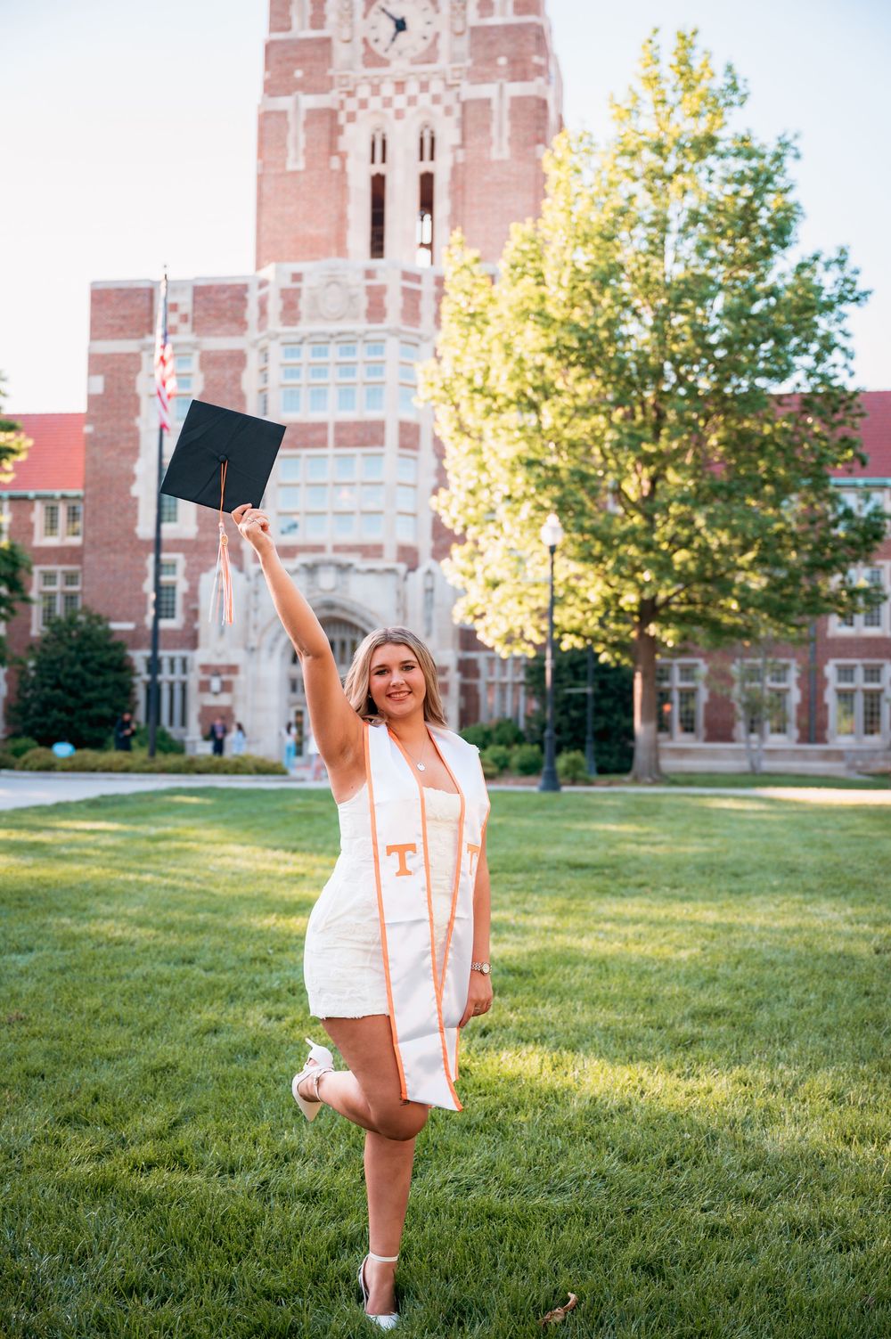 University of Tennessee graduate celebrates in white dress, raising cap in front of iconic brick campus building on sunny day.