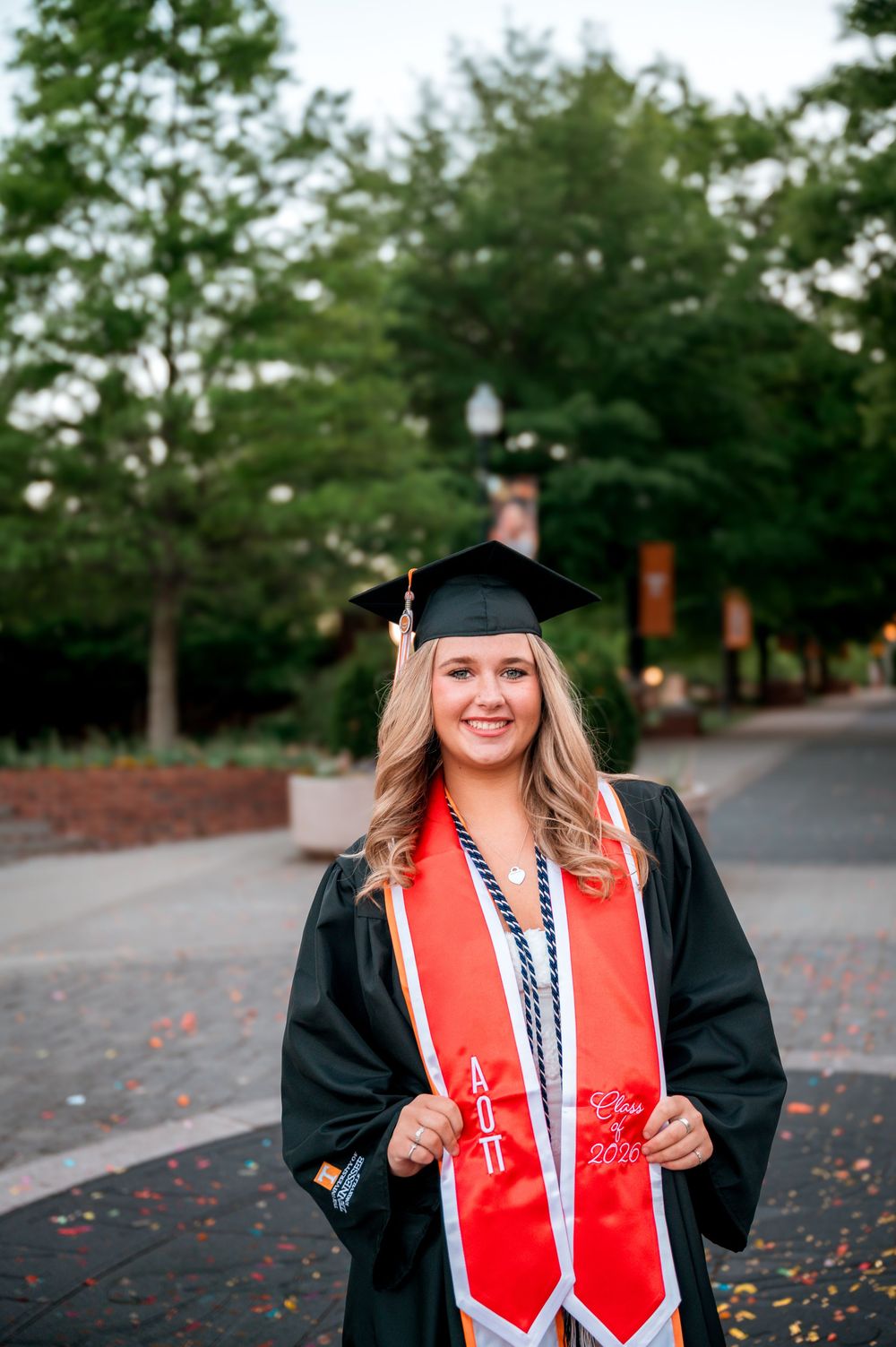 Smiling blonde woman in black graduation cap and gown with red AOII stole on campus.