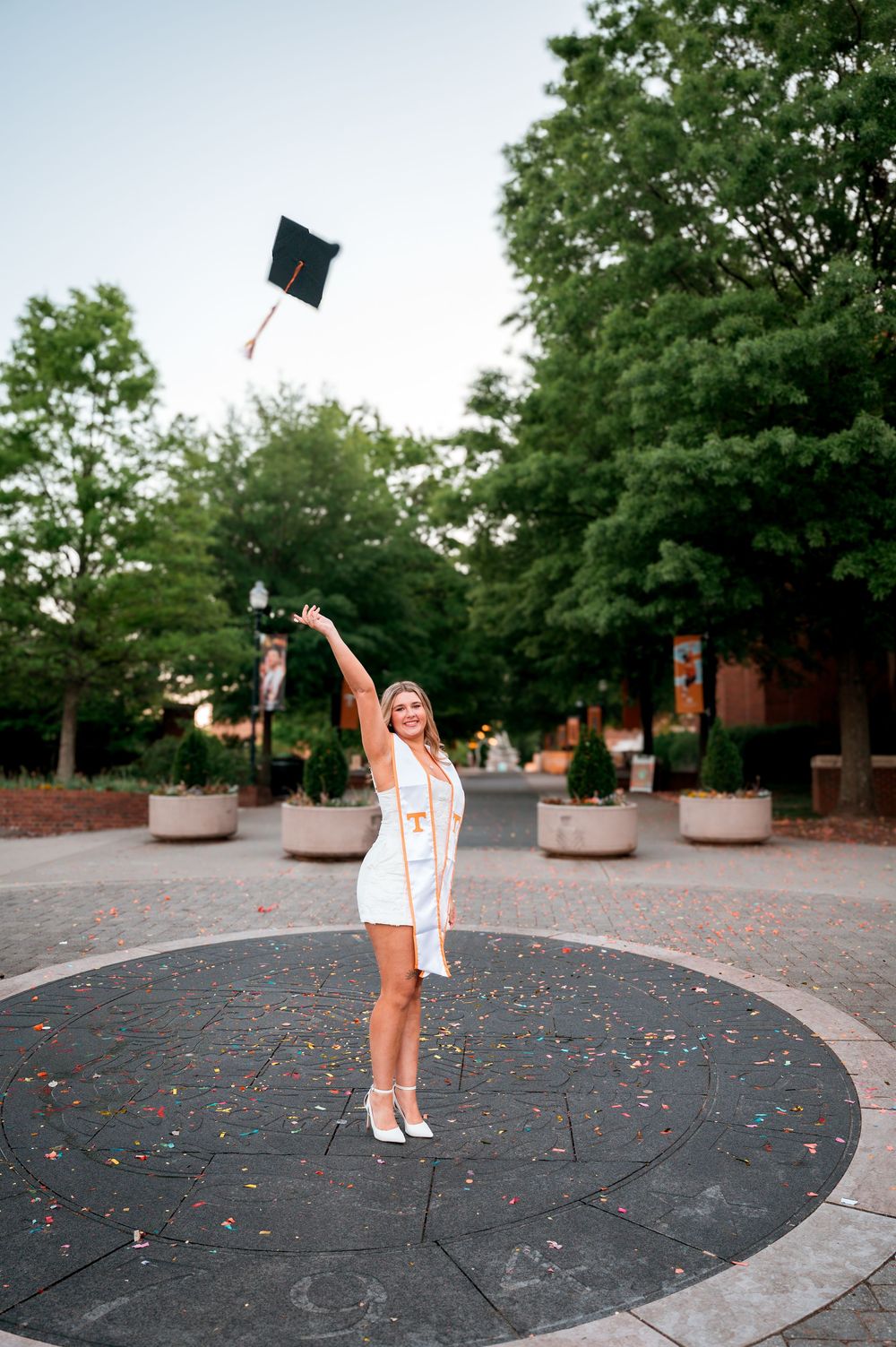 Smiling graduate in white dress tossing cap in the air outdoors on a decorative plaza surrounded by trees.