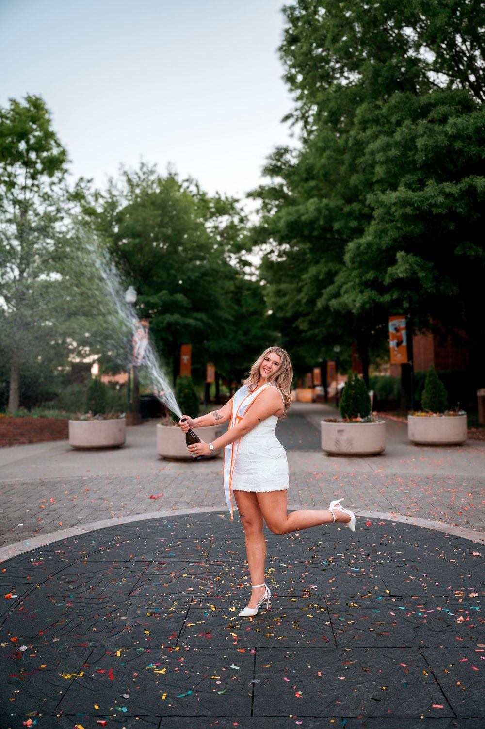 Joyful graduate in white dress popping champagne bottle on a colorful confetti-covered plaza with trees behind her.