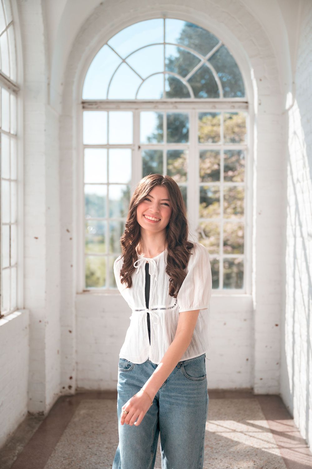 Smiling young woman in white blouse and jeans standing in sunlit room with arched windows.