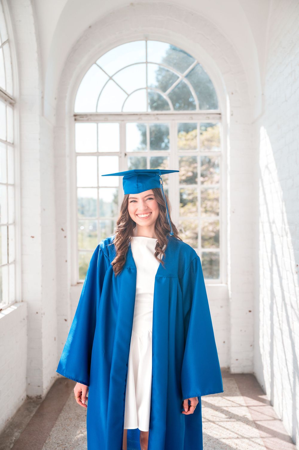 Young woman smiling in blue graduation cap and gown standing in bright white hallway with arched windows.