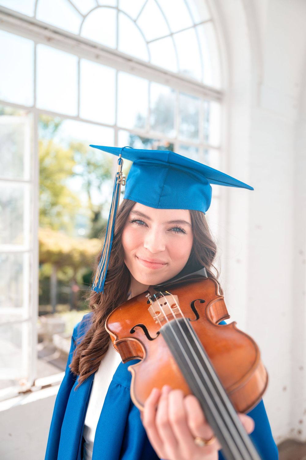 Young woman in blue graduation cap and gown smiling while holding a violin near a bright arched window.