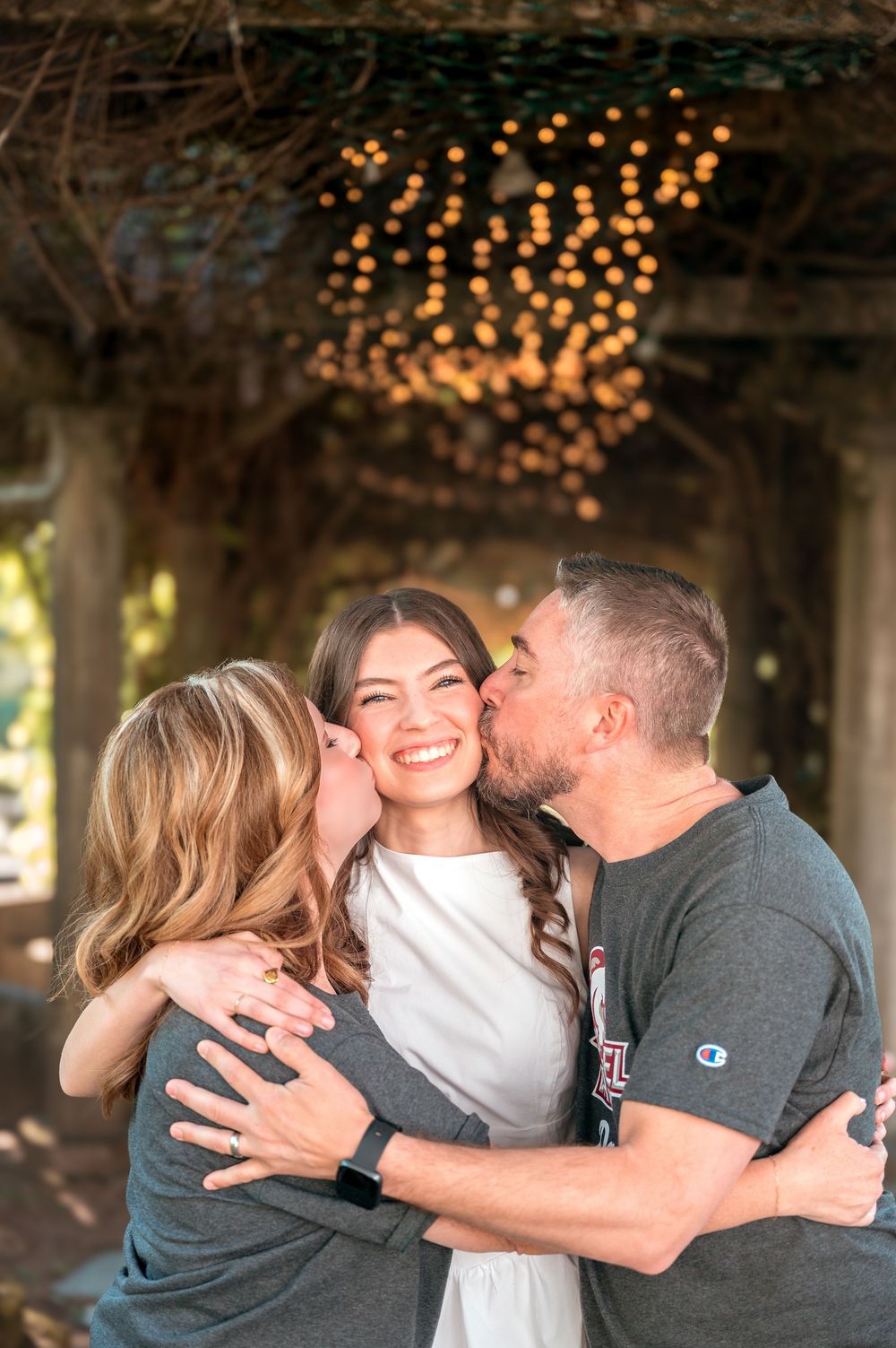 Parents kissing their smiling daughter on the cheeks during graduation celebration outdoors.