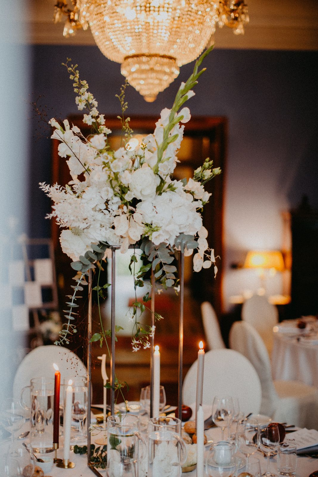 Elegant wedding reception table with tall white floral centerpiece and crystal chandelier overhead in dimly lit room.