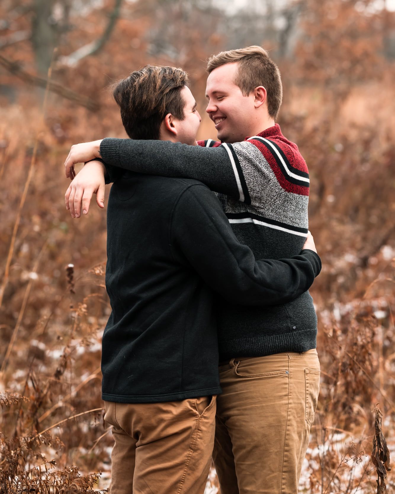 Two people embrace in an autumn field with dried grass and fallen leaves.