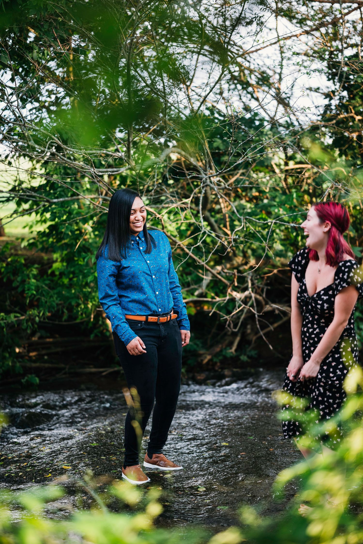 A couple shares playful moments by a stream in a lush green natural setting.