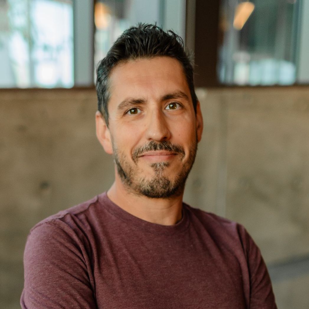 Professional headshot of person in burgundy t-shirt with facial hair smiling at camera in warm indoor setting.