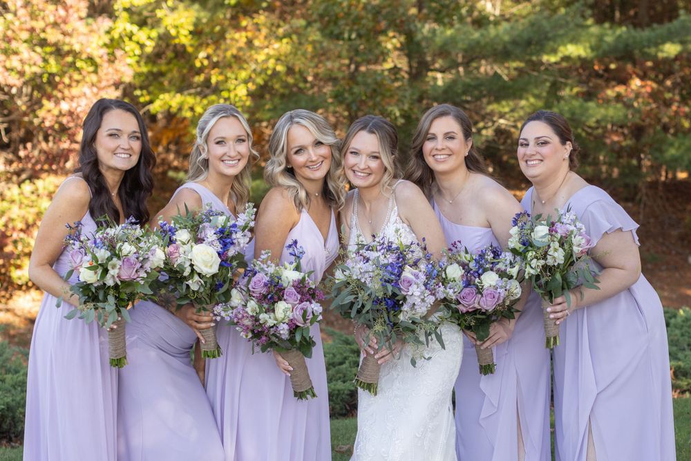 Bride with five bridesmaids in lavender dresses holding wildflower bouquets in autumn garden.