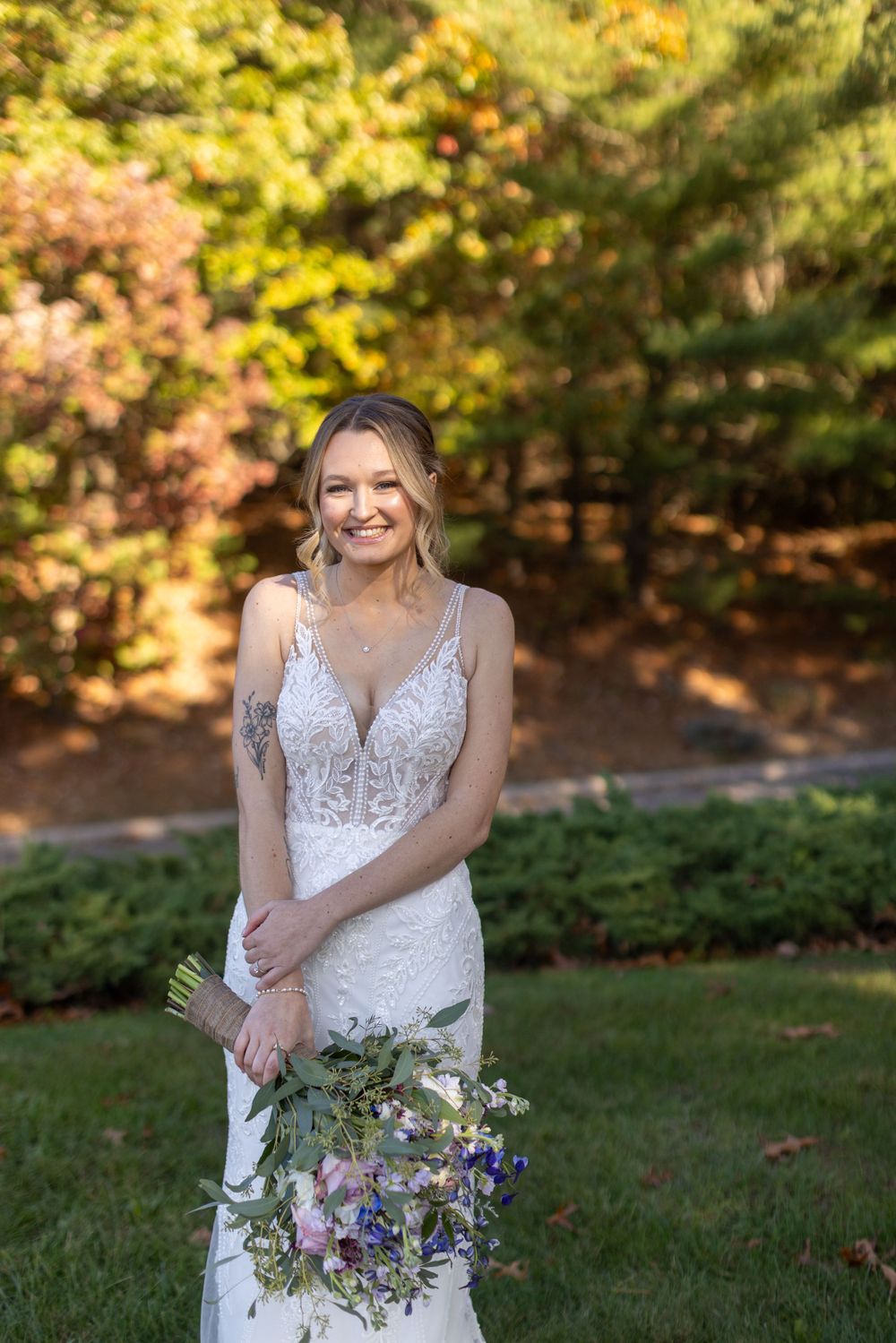Smiling bride in lace wedding gown holds wildflower bouquet in colorful autumn garden setting.
