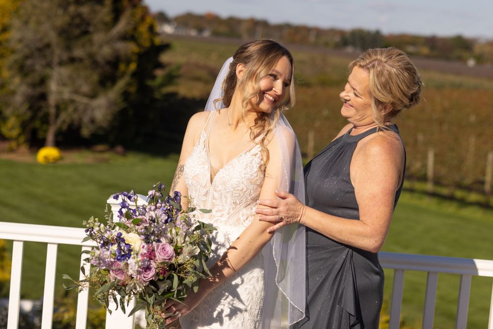 Bride in lace gown holding purple bouquet smiling with older woman in gray dress on outdoor deck.