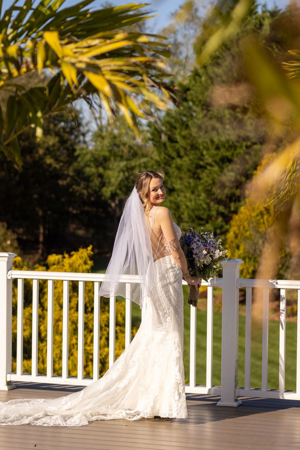 Bride in backless lace mermaid gown with veil posing on white railed deck surrounded by greenery.