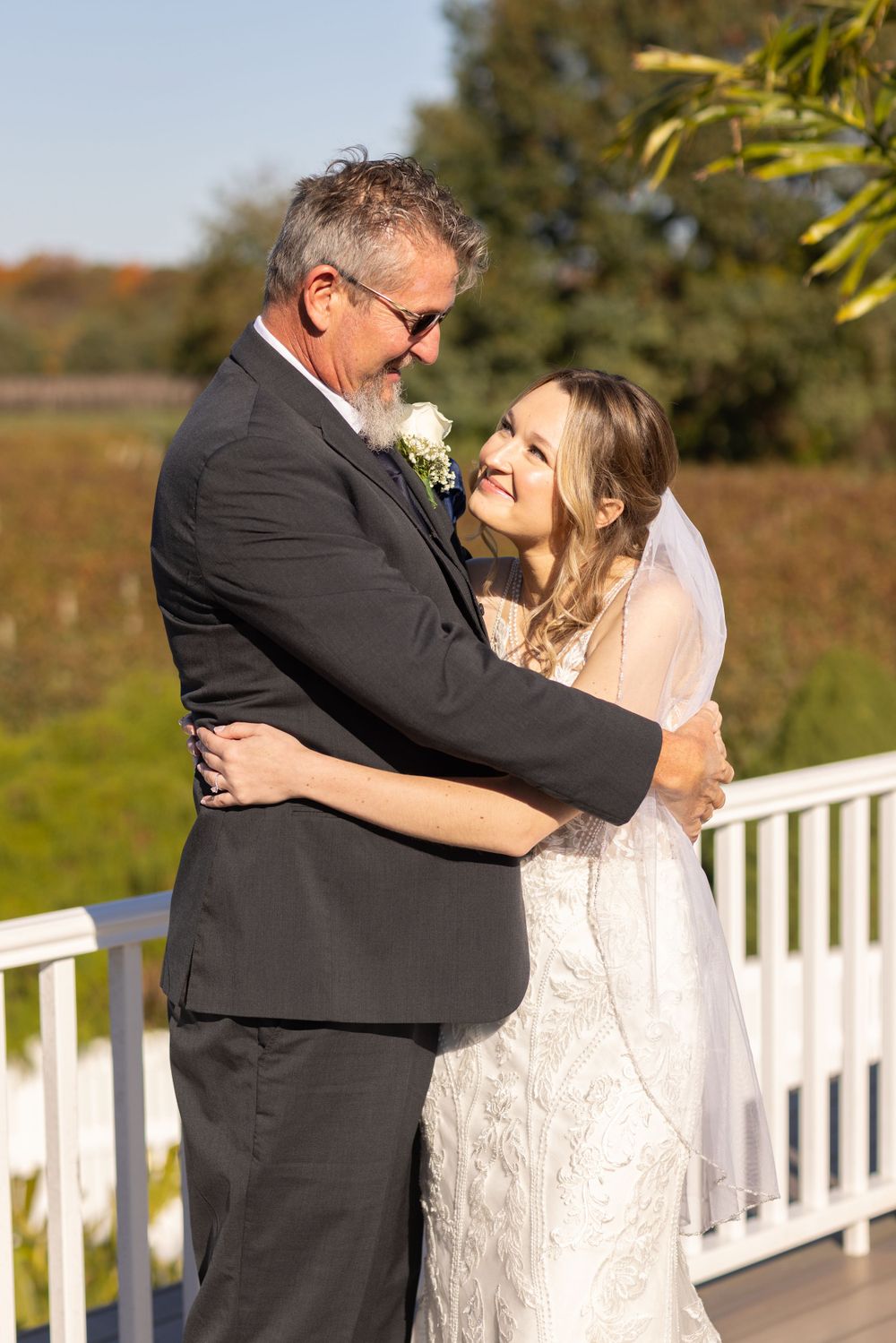 Bride in lace wedding dress embracing older man in gray suit on sunny outdoor deck at wedding.